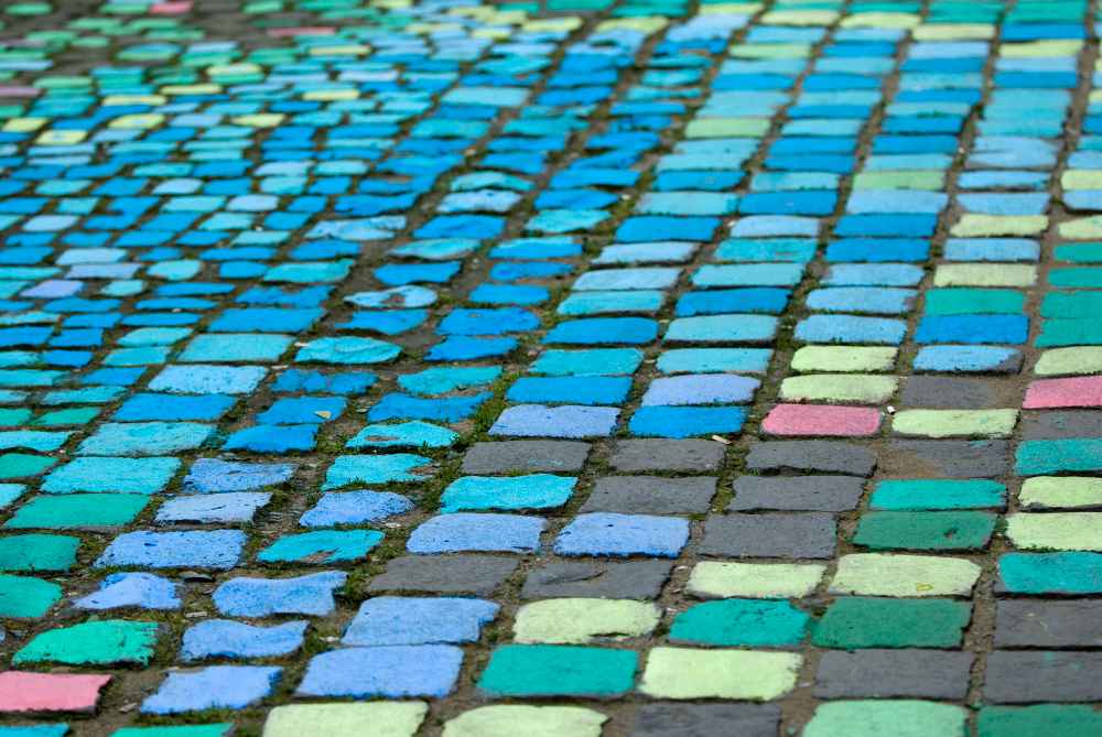 Allée pavée en grès naturels d'Inde colorés. Une vue en perspective d'une allée piétonne pavée de manière artistique. Les pavés, taillés dans des grès naturels d'Inde, ont été peints dans une palette de couleurs froides et vibrantes allant du bleu électrique au vert pomme, avec quelques touches discrètes de rose. Ce revêtement de sol original crée un effet de mosaïque urbaine texturée et moderne, capturant le regard grâce à ses nuances contrastées.