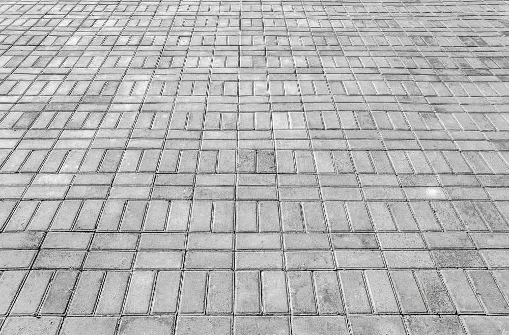 Une vue en perspective fuyante dévoilant une vaste surface pavée à l'extérieur. Le sol est composé de dalles rectangulaires de Kandla gris, une pierre naturelle réputée pour sa robustesse et ses nuances minérales subtiles. Les blocs sont agencés selon un motif géométrique rigoureux alternant des sections horizontales et verticales, ce qui crée un rythme visuel structuré et contemporain. La texture légèrement texturée de la pierre accroche la lumière, offrant un dégradé de gris clair à gris moyen qui renforce l'aspect épuré et design de cet aménagement urbain ou paysager.