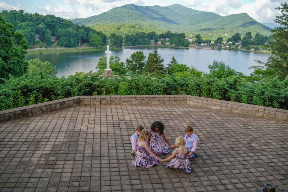 Terrasse en pavé grès Kandla avec vue sur le lac. Cinq enfants en tenues de fête sont assis en cercle au centre d'une vaste esplanade extérieure. Le sol de cette terrasse est entièrement revêtu de pavé grès Kandla aux nuances ocres et grises, offrant une surface stable et authentique. En arrière-plan, une murette en pierre délimite l'espace avant de laisser place à une végétation dense, une grande croix blanche, un lac paisible et des montagnes verdoyantes sous un ciel légèrement nuageux.