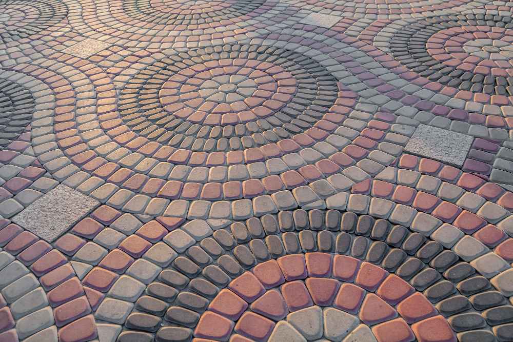Pavés Kandla : élégance et design en motifs circulaires. Cette image montre en détail une surface pavée en pavés Kandla naturels. Les pierres sont disposées selon un motif circulaire complexe et attrayant, créant un effet visuel de cercles concentriques qui se chevauchent. La palette de couleurs naturelles des pavés Kandla, allant du gris moyen au rose terreux et au rouge chaud, renforce l'esthétique rustique et authentique de ce pavage. Des carreaux plus clairs sont également visibles, ajoutant du contraste et des détails architecturaux au design global. Ce type de pavage est idéal pour créer des espaces extérieurs durables et élégants, comme des allées, des patios ou des places publiques.